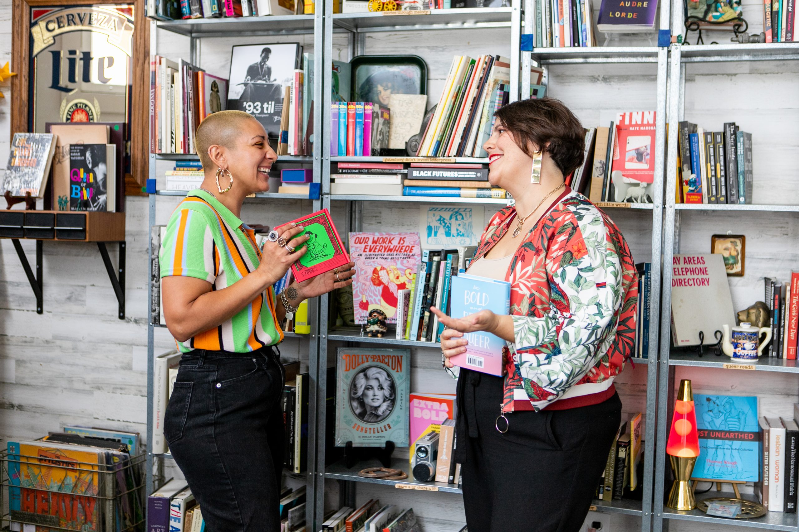 Two queer people talk in a bookstore in front of a shelf of books. One on the right has a shaved head that's died yellow and a fun lime green shirt on. The other on the right is wearing a floral pattern jacket with a book in her hand. Both are talking to each other.